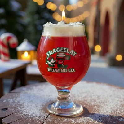 Red beer glass with white foam and a lit candle on top, featuring Frageelay Brewing Co. logo, outdoors with blurred lights and Christmas decorations.